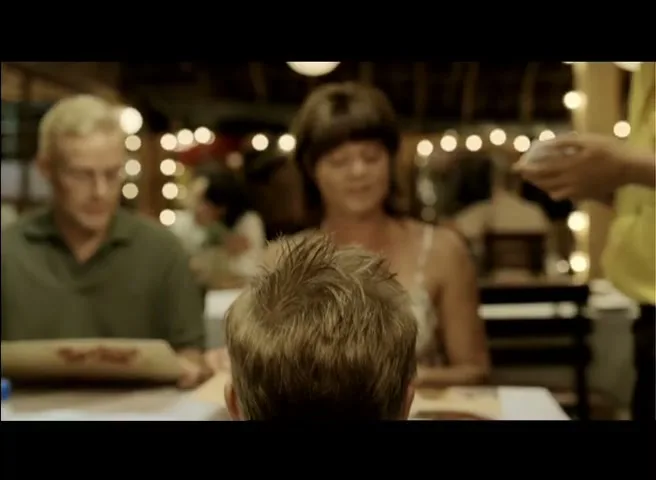 A young boy with spiky blonde hair sits at a restaurant table, with his parents and a server in the background.