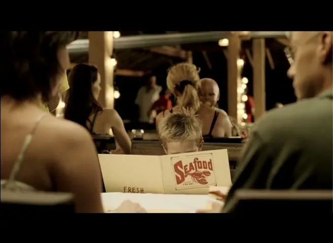 A young boy with blonde hair peeks over a "Seafood" menu at a restaurant table, surrounded by adults at night.