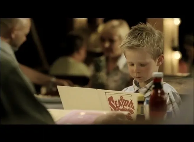 A young boy with blonde hair sits at a table in a dimly lit restaurant, looking at a "Seafood" menu.