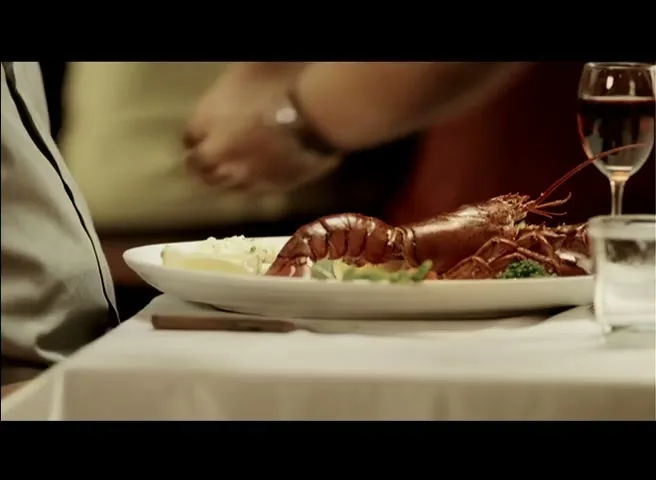 A waiter's hands place a plate with a cooked lobster on a white tablecloth in a restaurant, with a glass of red wine visible.