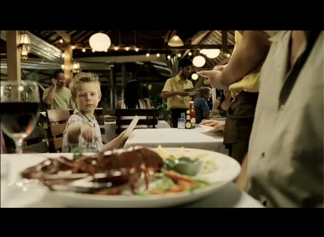 A young boy in a plaid shirt sits at a restaurant table, looking at the camera, with a plate of lobster in the foreground and other diners in the background.