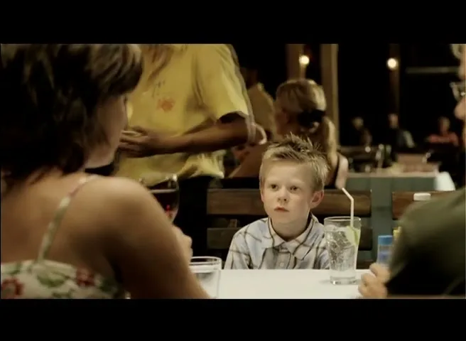 A young boy with spiky blonde hair sits at a restaurant table between two adults, looking around with a curious expression at night.