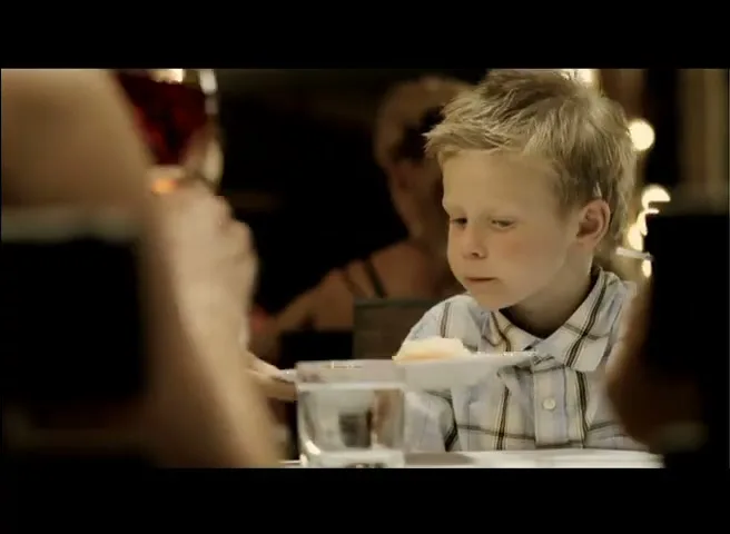 A young boy with blonde hair sits at a restaurant table, looking down as a plate is placed in front of him.