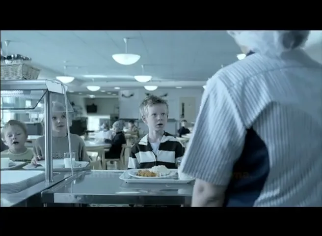 A young boy in a striped shirt looks up at a cafeteria worker while holding a tray of food in a school cafeteria.