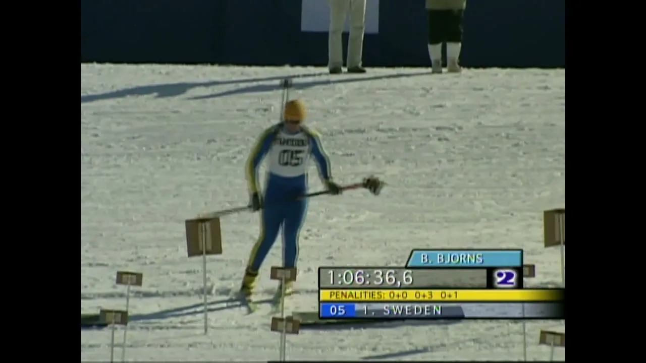 A male biathlete in a blue and yellow suit skis towards the shooting range, preparing to aim his rifle at targets in a snowy, outdoor setting.