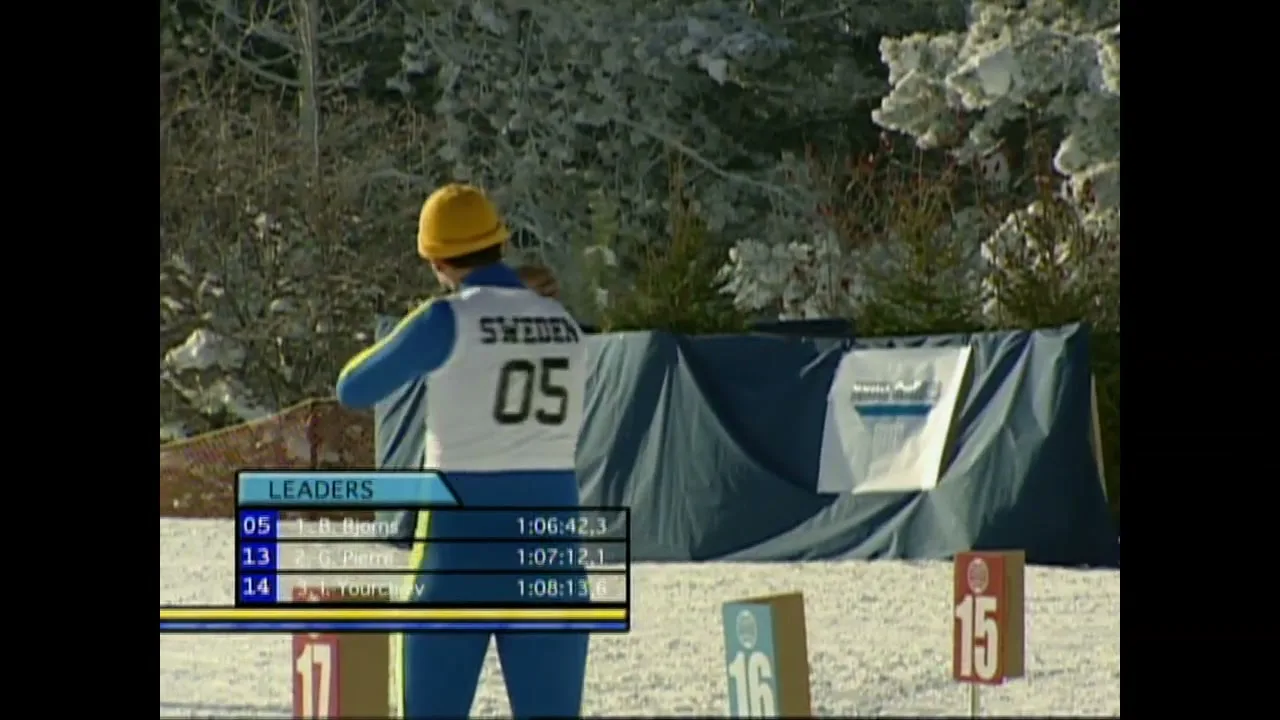A male biathlete in a blue and yellow suit with number 05 on his back aims a rifle at a target in a snowy, wooded area.