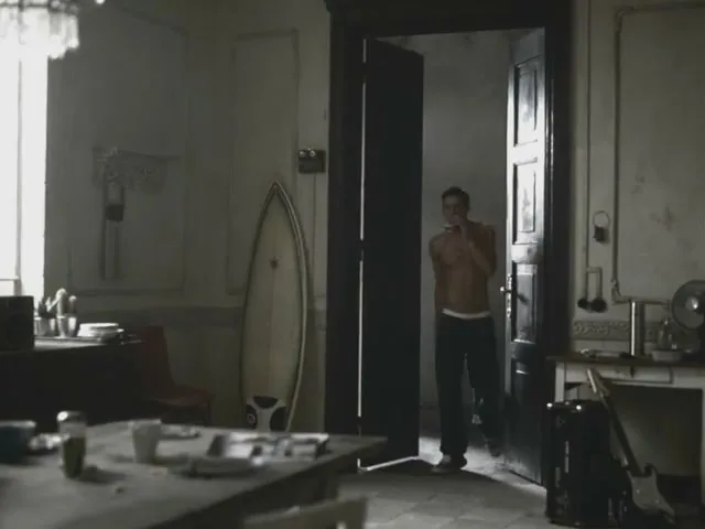 A shirtless man in dark pants walks through a doorway into a messy, dimly lit room with a surfboard leaning against the wall.