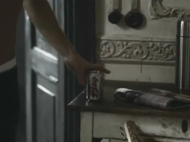 A shirtless man drinks from a can of Coca-Cola Light, then places it on a rustic wooden table in a kitchen.