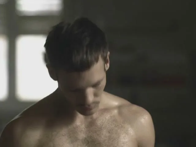 A shirtless, sweaty man with a mustache looks at the camera, then down, then back up, in a dimly lit room.