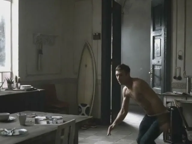 A shirtless man in dark pants turns to run away from a woman in underwear drinking a Coca-Cola Light in a dimly lit apartment.