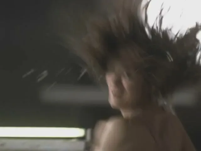 A woman with long brown hair, wearing a bra, is seen from behind, turning her head and smiling, with her hair flying up in a dark room.