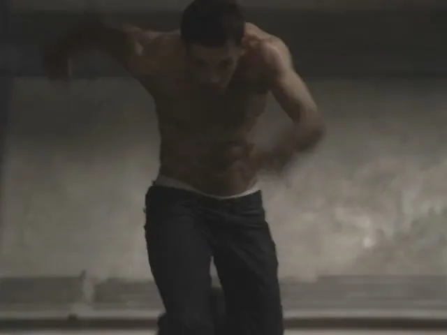 A shirtless man in dark pants runs towards the camera in a dimly lit, industrial-looking interior space.