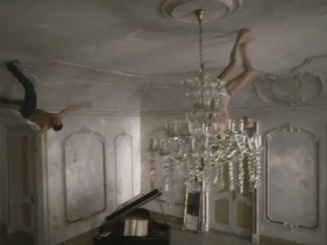 A man and a woman, both shirtless, hang upside down from the ceiling of a grand, ornate room, reaching for each other.