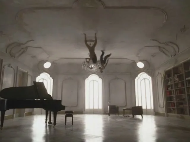 Two men in casual clothes are hanging upside down from the ceiling of a grand, ornate room with a piano and large windows.