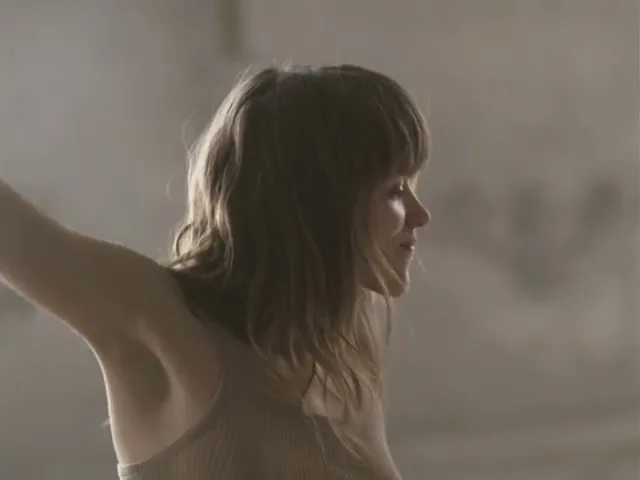 A young woman with light brown hair and a beige tank top smiles and looks to the right, with her arm outstretched in a bright, airy room.