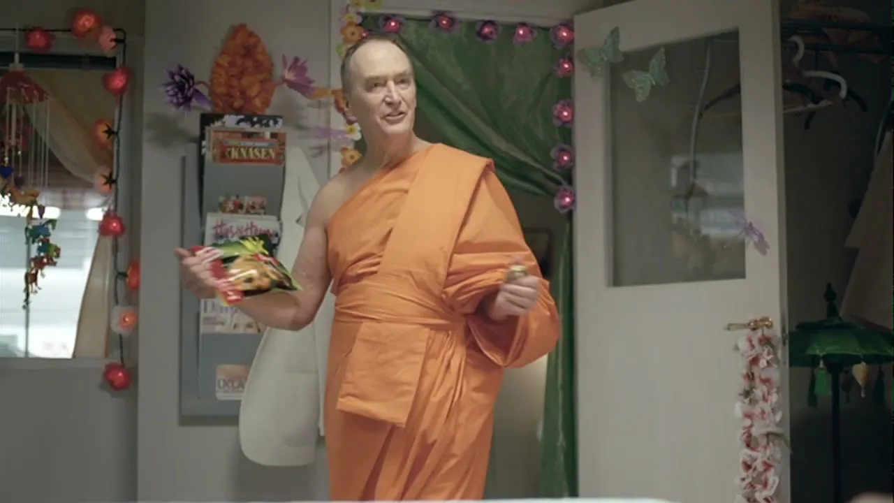 A middle-aged man dressed as a Buddhist monk stands in a colorful room, holding a small bag of chips and a bell, looking around.