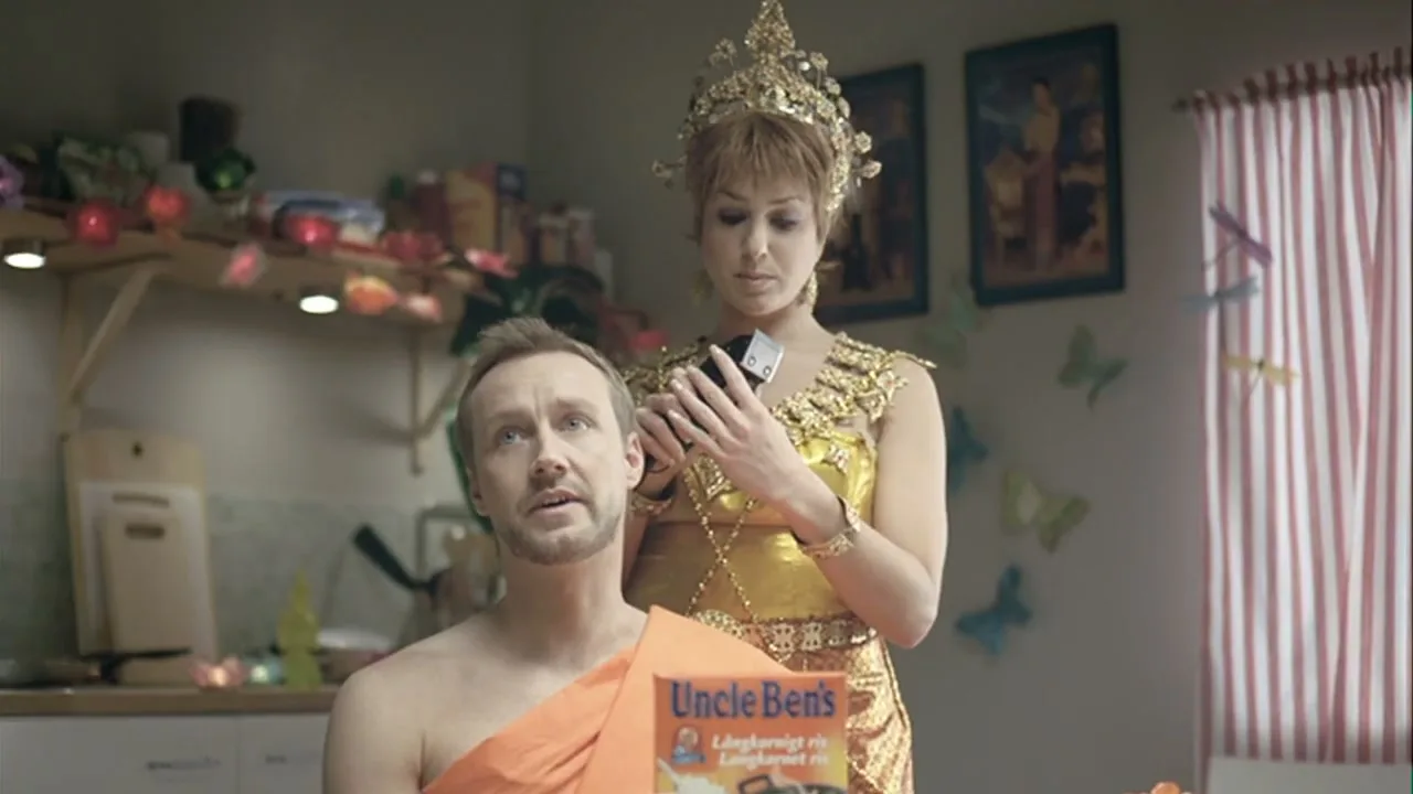 A man dressed as a monk sits with a woman in traditional Thai attire behind him, holding a hair clipper, in a kitchen.