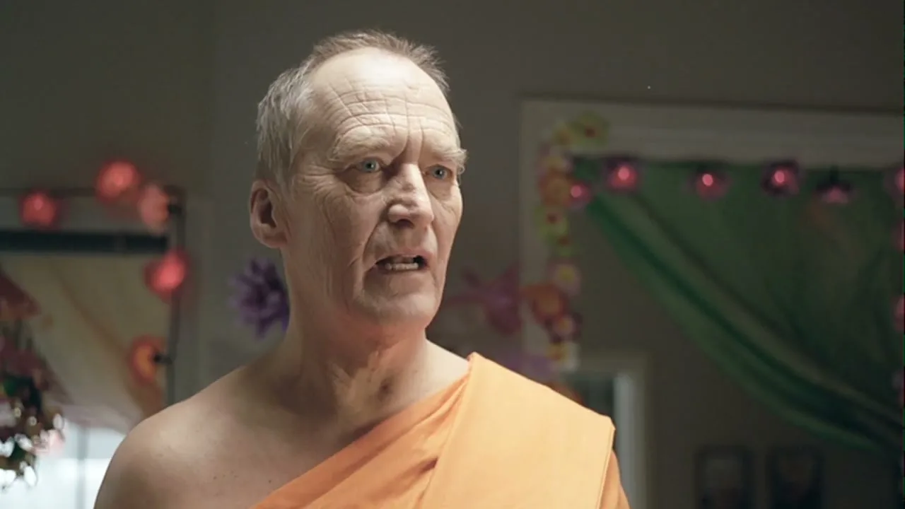 An older man with a shaved head and an orange robe looks around a room decorated with colorful paper lanterns and garlands.