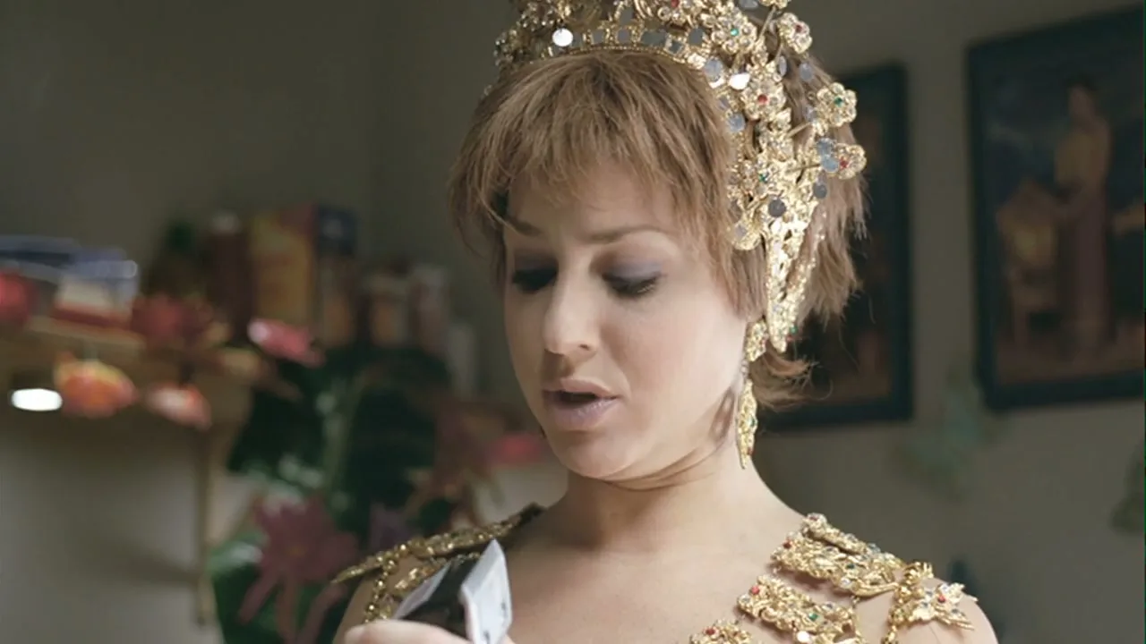 A woman in her 30s, dressed in an elaborate gold costume and crown, looks down at a small object in her hand with a surprised expression in an interior setting.