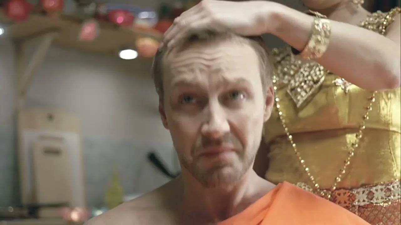 A man in an orange robe looks up in fear as a woman in traditional Thai clothing touches his head in a kitchen.
