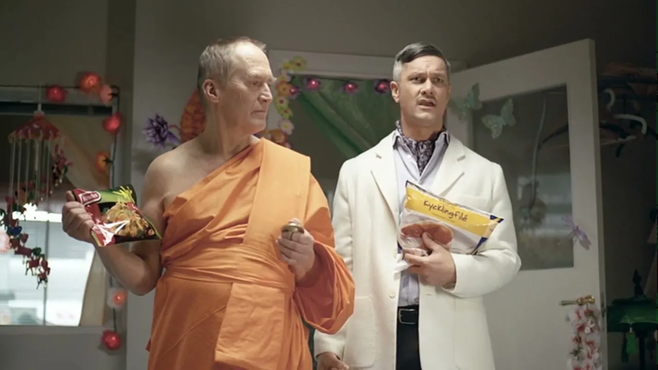 Two men, one dressed as a monk and the other in a white suit, stand in a decorated room, holding grocery items and looking at each other.