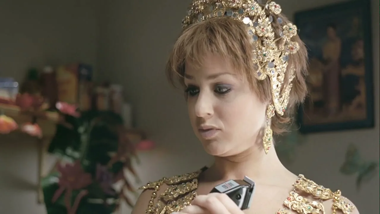 A woman in a golden, ornate costume and crown holds an electric shaver to her neck, looking concerned in a domestic interior.