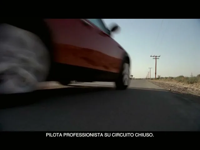 A red Mini Cooper drives past a "Road Closed Ahead" sign on a desert road under a clear sky.