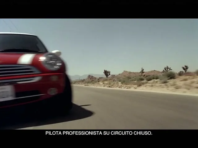 A red and white Mini Cooper drives quickly down a desert road under a clear blue sky, with mountains and sparse vegetation in the background.