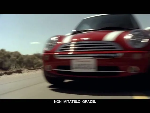 A red Mini Cooper with white racing stripes drives quickly on a desert road under a clear blue sky, with scrub brush visible on the left.