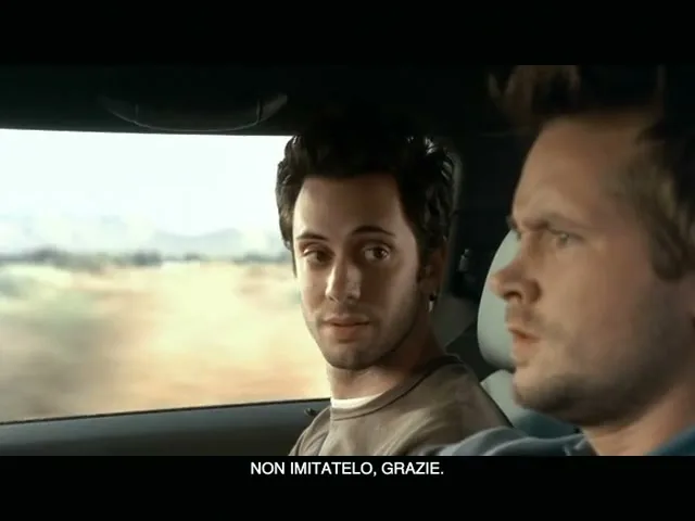 Two men in a car, one looks at the camera, the other looks forward, as they drive through a desert landscape.