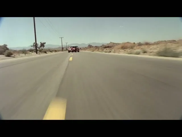 A red Mini Cooper drives away from the camera down a long, straight desert road under a clear blue sky.