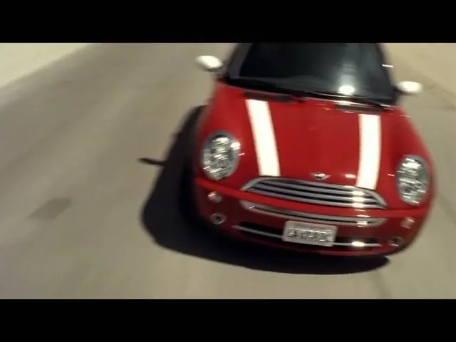 A red Mini Cooper with white racing stripes drives on a desert road under a bright sky.