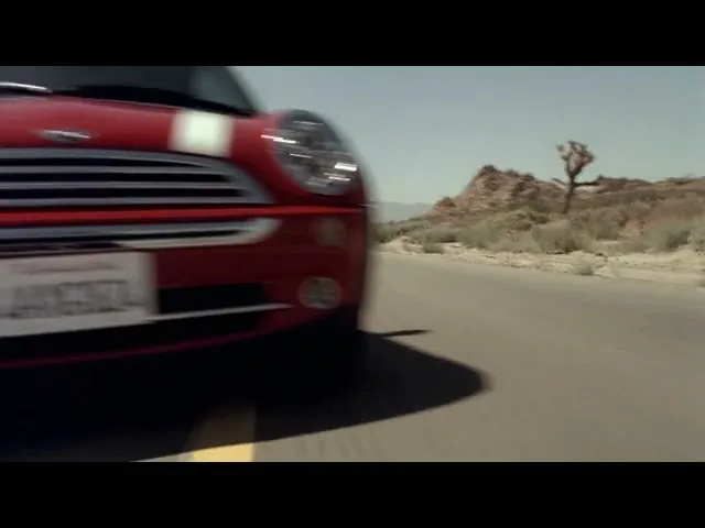 A red Mini Cooper with white racing stripes drives quickly on a desert road, with mountains and a Joshua tree in the background.