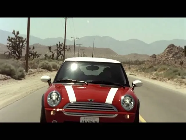 A red Mini Cooper with white racing stripes drives towards the camera on a desert road under a bright sun.
