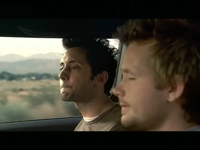 Two men in their 30s are driving in a car, looking out the window at the passing landscape during the day.