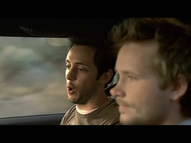 Two men in a car, one with dark hair and a brown shirt, the other with blonde hair, are driving and talking.
