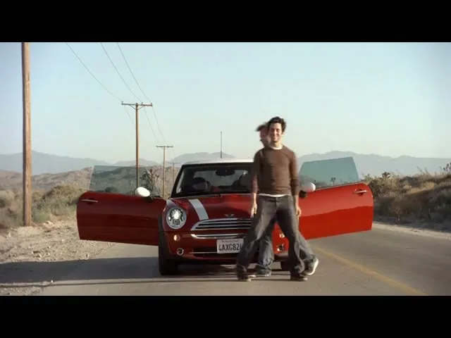 Two men in casual clothes get out of a red Mini Cooper on a desert road with mountains in the background under a clear sky.