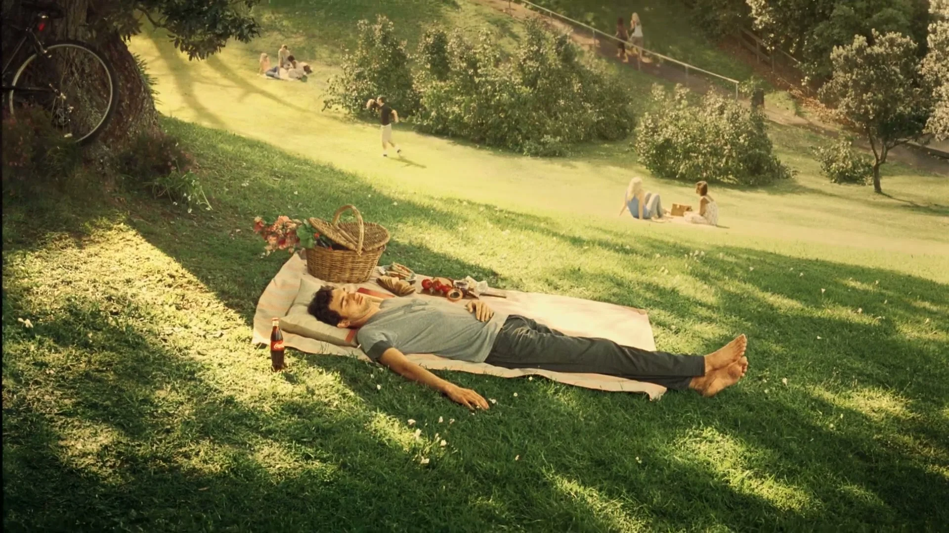 A man in a grey t-shirt and dark pants lies barefoot on a picnic blanket under a tree in a sunny park, with other people in the background.