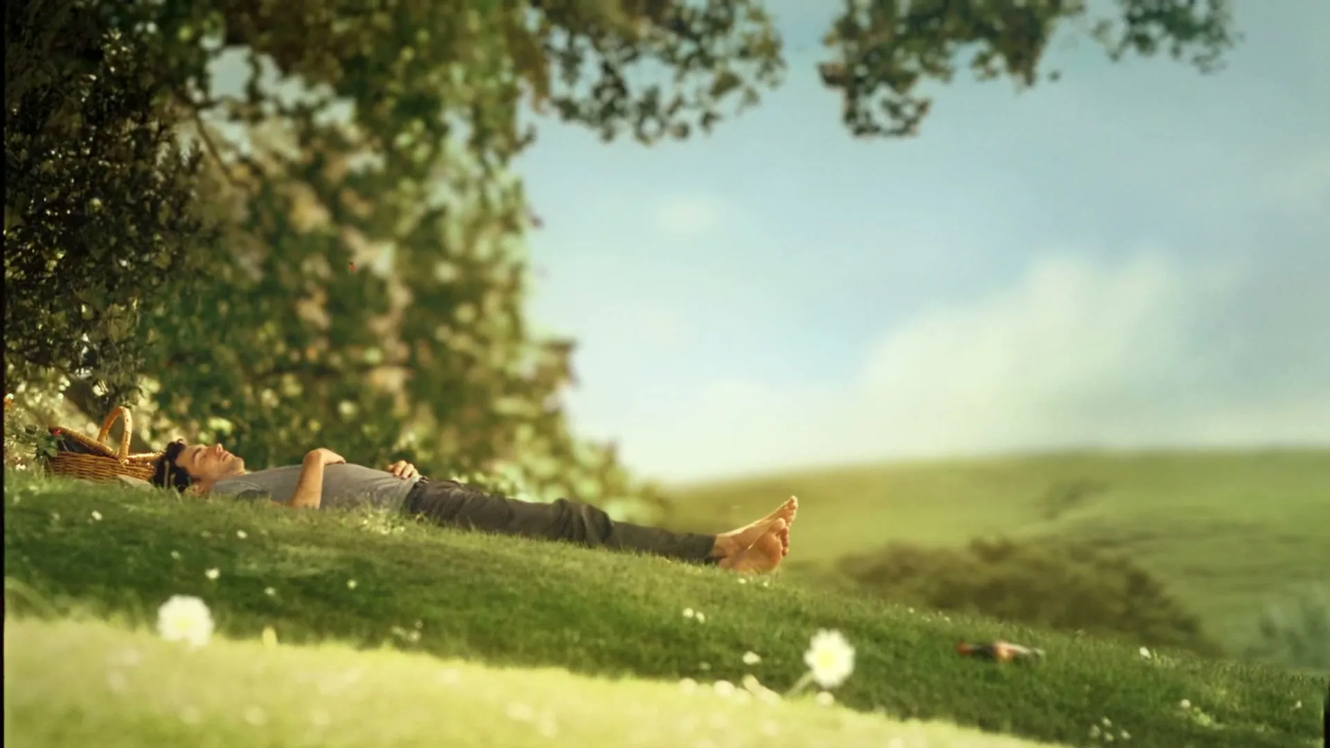 A man in a grey t-shirt and dark pants lies barefoot on a grassy hill under a tree, with a picnic basket nearby, enjoying a sunny day.