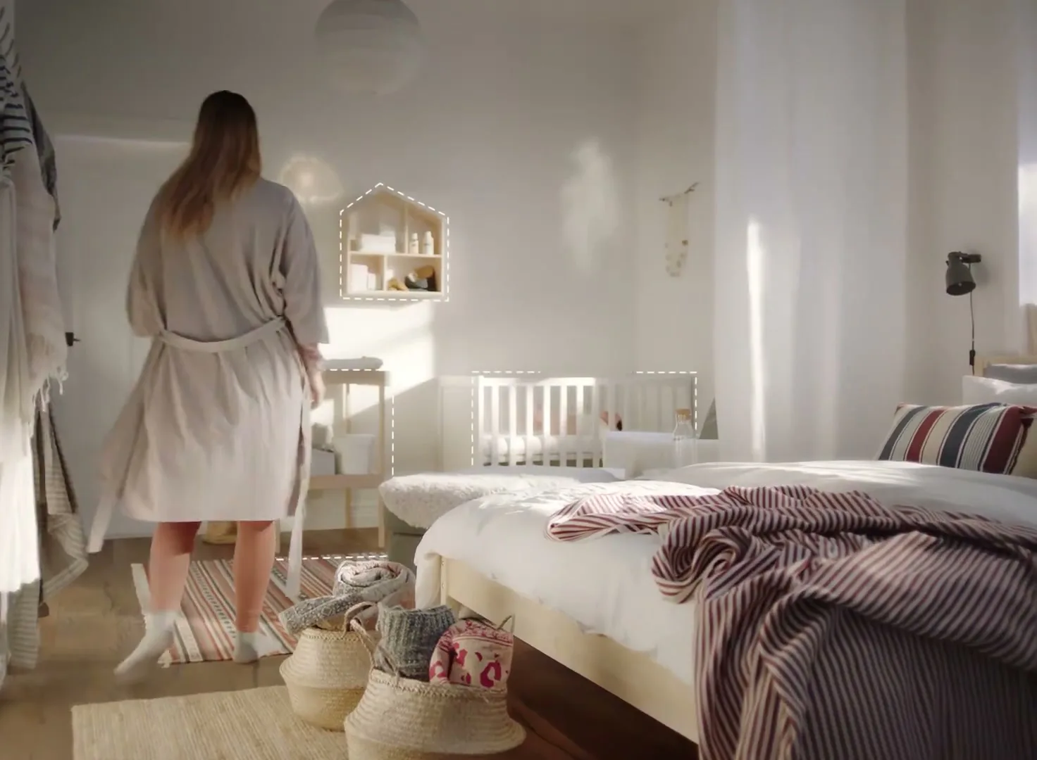 A woman in a robe walks through a sunlit bedroom, past a bed and towards a baby's crib, with baskets on the floor.