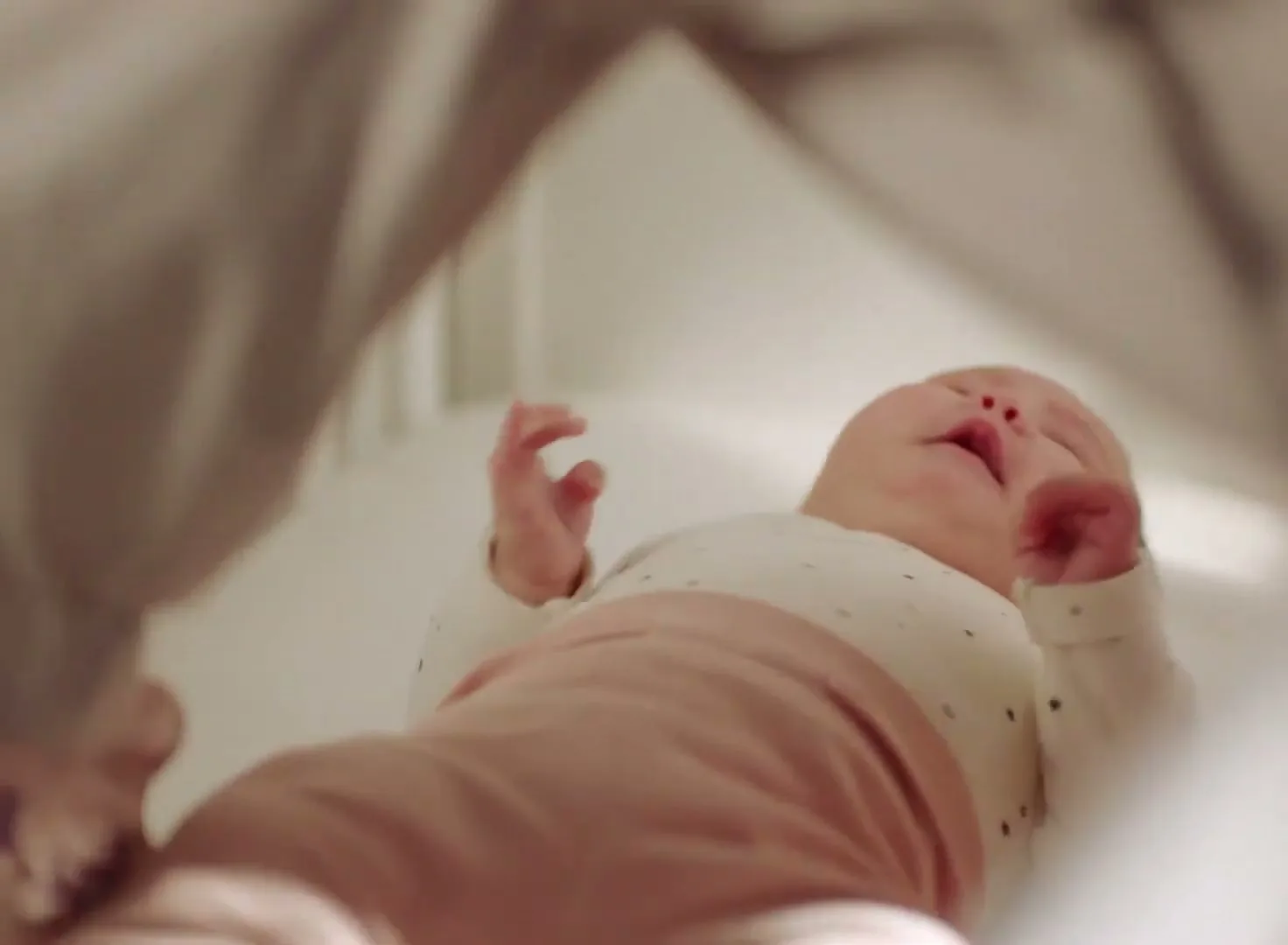 A baby in a white onesie and pink pants lies in a white crib, crying, as an adult's hand reaches in to comfort it.