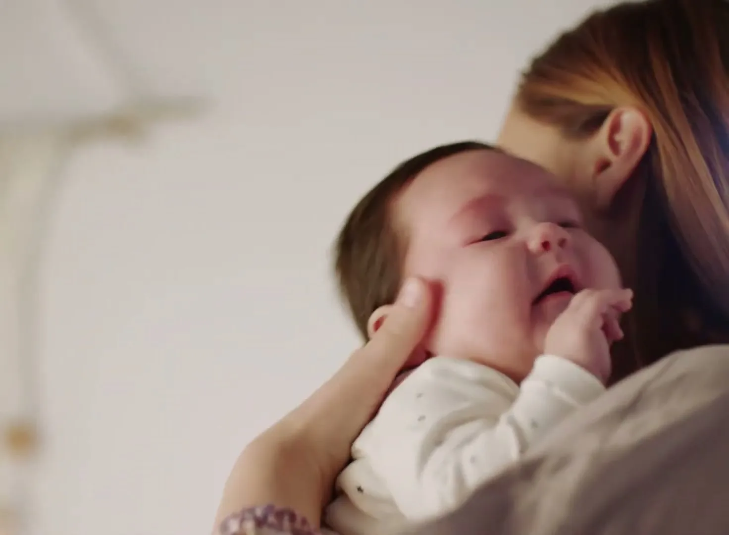 A mother with long brown hair holds her baby, who is wearing a white onesie, over her shoulder in a brightly lit room.