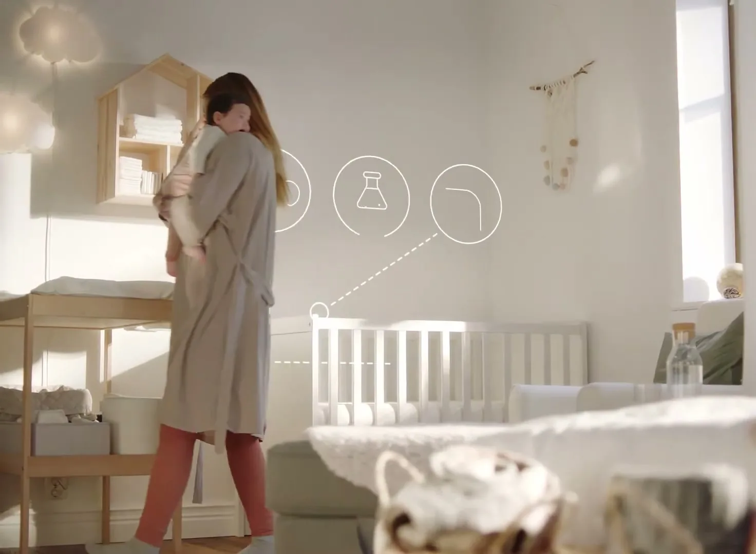 A mother in a robe holds her baby, then places the baby on a changing table in a bright nursery with a crib and shelves.