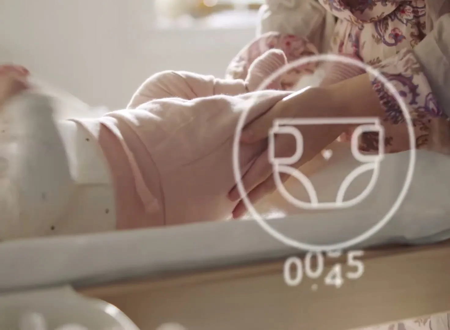 A baby in a white onesie and pink pants lies on a changing table while an adult's hands change its diaper, with a graphic overlay of a diaper and numbers.