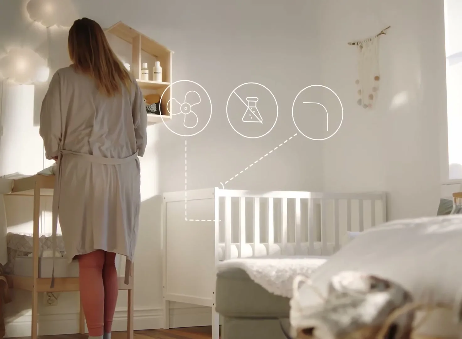 A woman in a robe changes a baby's diaper on a changing table in a bright, white nursery, with a crib and bed in the background.