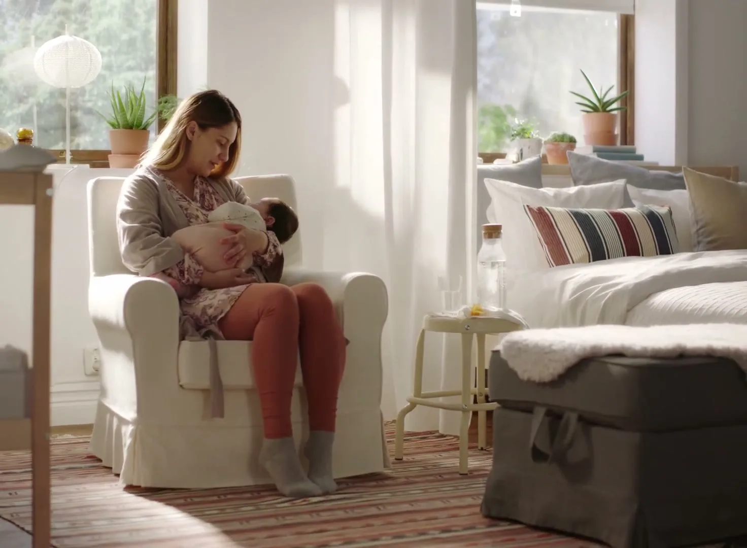A young mother sits in a white armchair, cradling her newborn baby in a sunlit bedroom, looking down at the baby with a gentle smile.