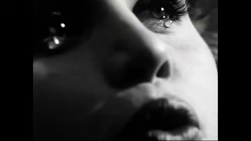 A woman's face is shown in extreme closeup, with a tear visible in her eye, as she whispers in a dramatic black and white shot.