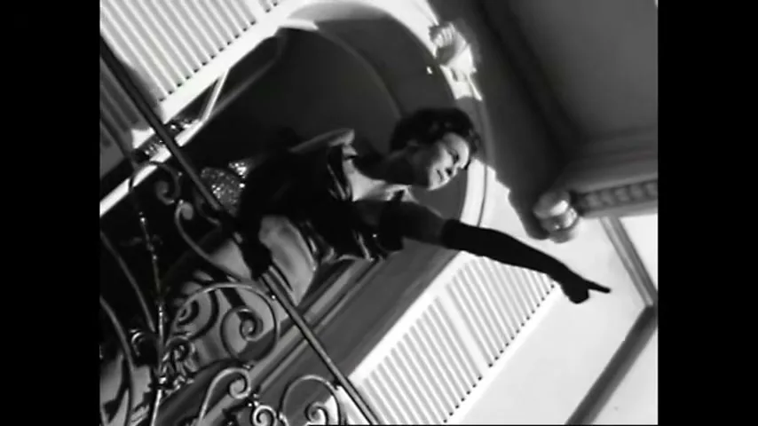 A woman in a strapless dress and long gloves leans over a balcony, pointing and shouting in a dramatic, low-angle shot.