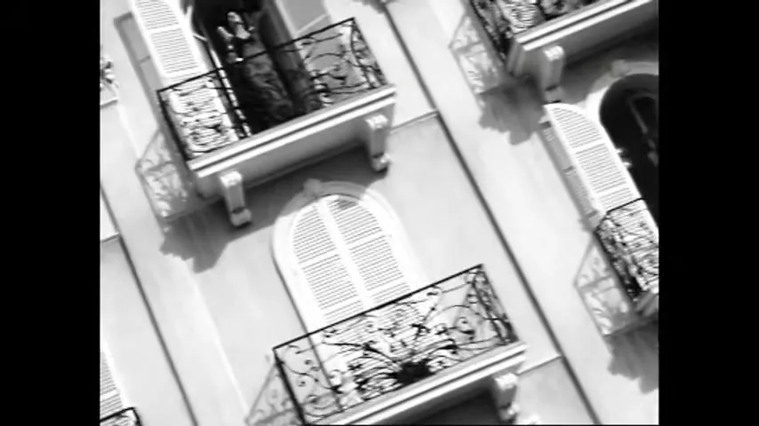 A black and white shot of a building with multiple balconies, where women in dark dresses are seen shouting and gesturing from their windows.