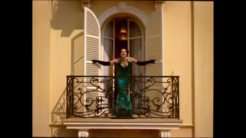 A woman in a green dress and black gloves stands on a balcony, arms outstretched, shouting at an unseen person.
